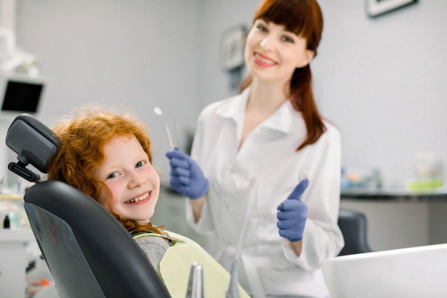 Smiling red-haired child in a dental chair with a friendly female dentist giving a thumbs up
